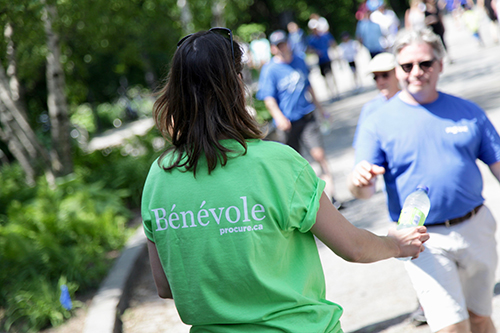 Bénévole vue de dos tendant une bouteille de H2O à un participant de la Marche du Courage PROCURE